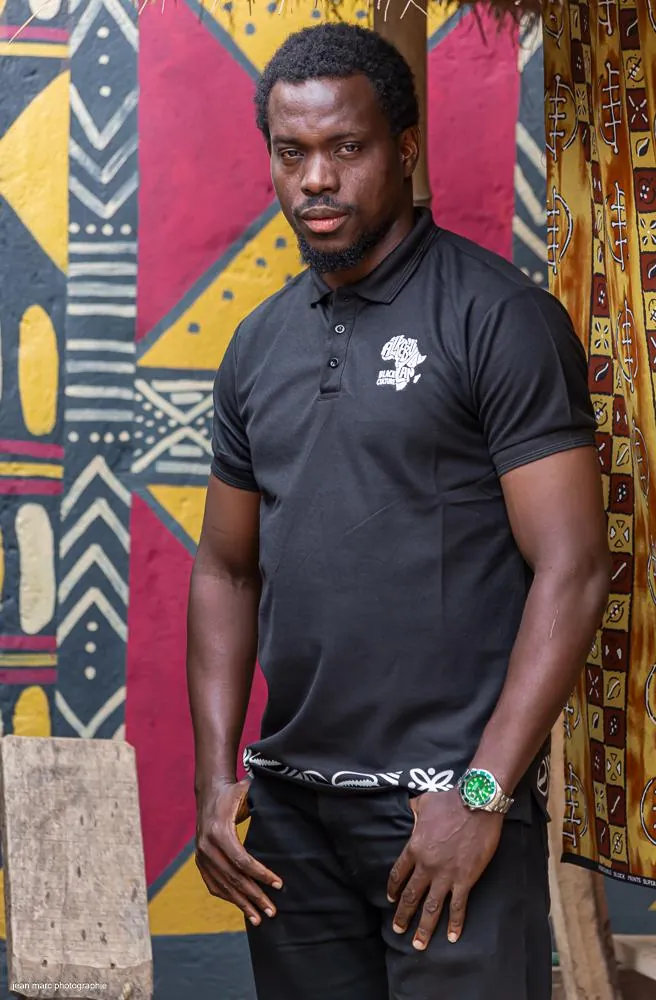 Homme en polo noir Alkebulan devant un mur peint de motifs africains
